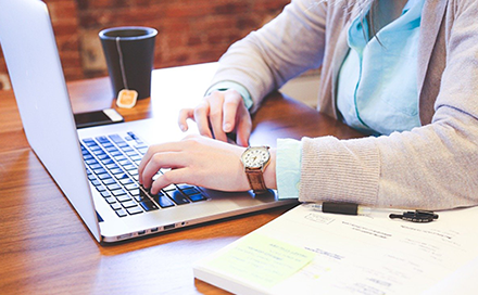 A person works on a laptop with a notebook and tea