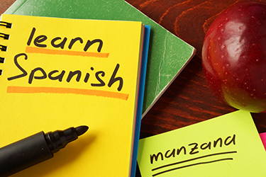 Notebook titled "Learn Spanish" next to an apple with a piece of paper below it with "manzana" written on it.