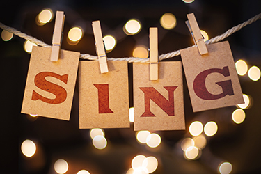 Each letter of the word "SING" is cut out and attached to a rope with a clothes pin with soft lights in the background.