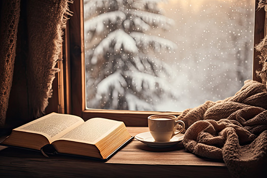 An open book next to a cup of tea in front of a window looking out at a snowy tree.